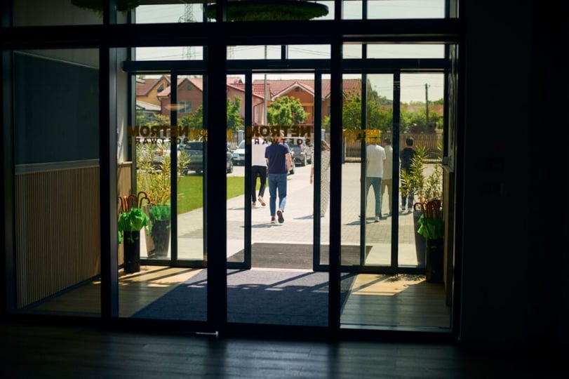 entrance.webp Mensen lopen door de glazen ingang van de NetRom Campus.