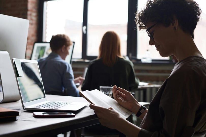 Persoon leest notities naast een laptop terwijl collega’s op de achtergrond werken.